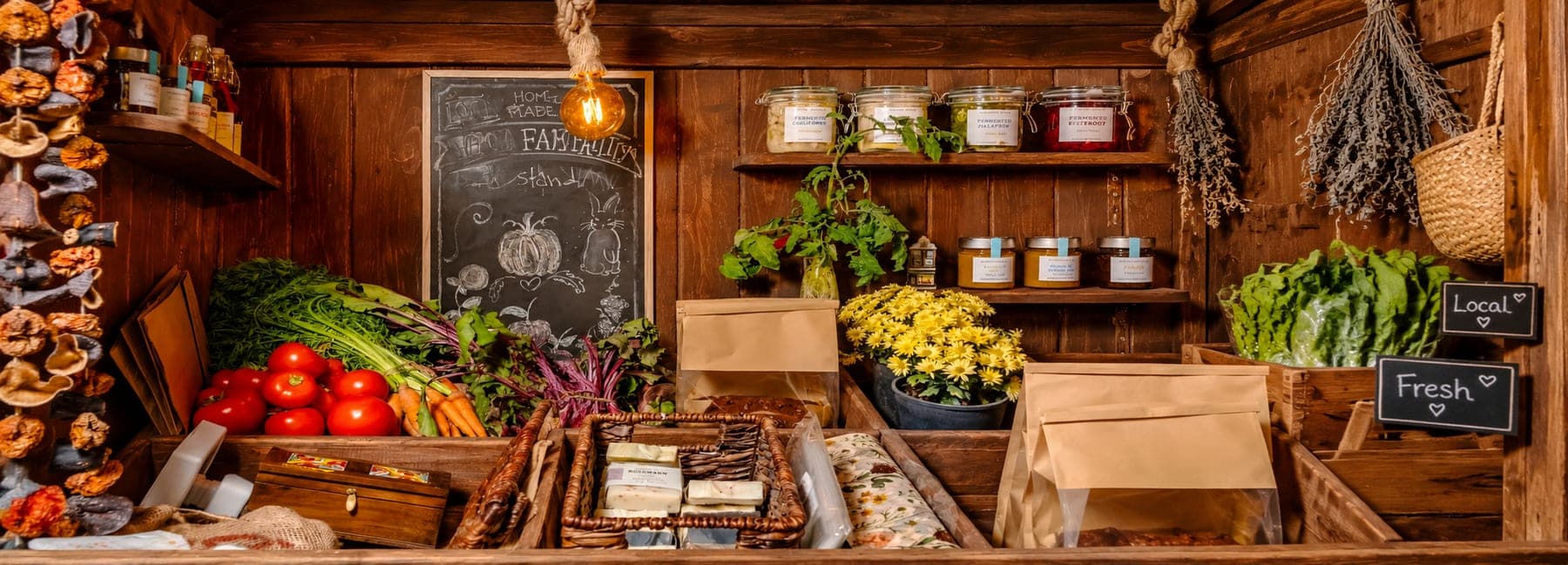 A farm stand of tomatoes, carrots and beet greens, with jars of preserves on wooden shelves, baskets of soap and a chalkboard sign